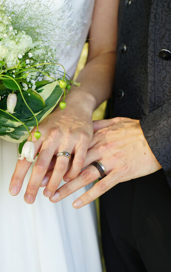 Hände von einem Hochzeitspaar. Eheringe sind zu sehen. Die Braut hält einen Strauß in der rechten Hand.