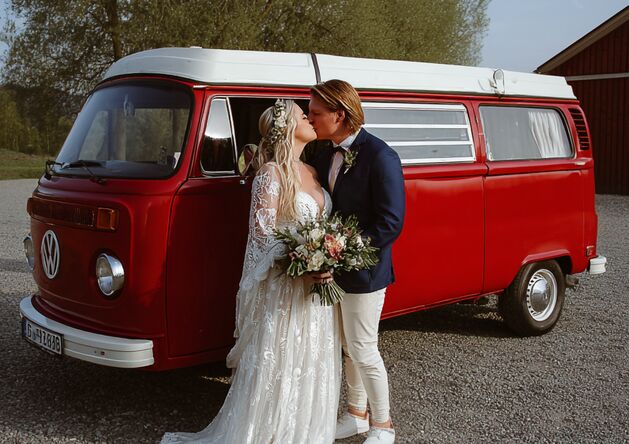 Hochzeitspaar vor altem, roten VW-Bus. Baum im Hintergrund.