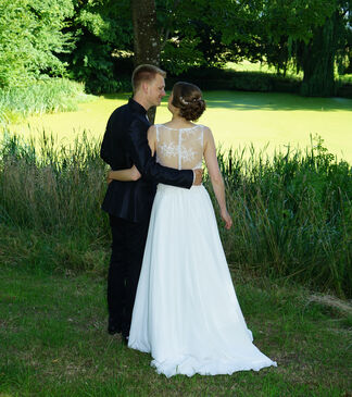 Hochzeitspaar an einem Seeufer. Paar steht mit Rücken zum Betrachter. Bäume im Hintergrund.